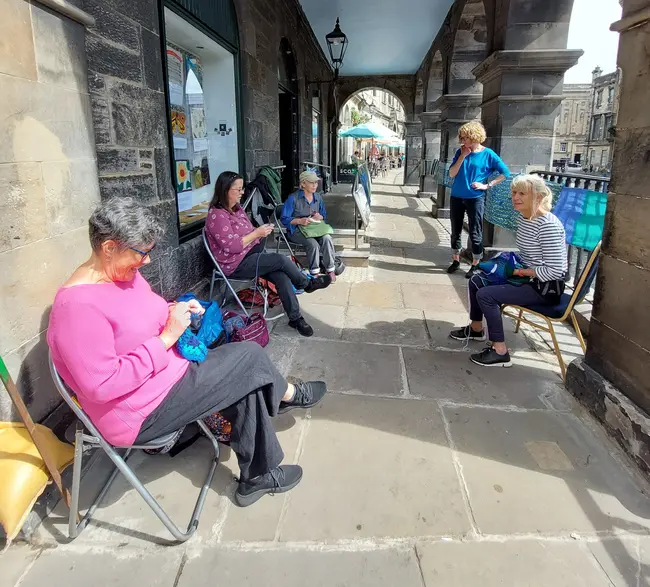 A socially distanced group of people sit outside a Meeting House. They are crafting embroidered panels. You can see more panels in the window of the Meeting House behind them. They look friendly and approachable. 
